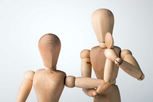 Symbolbild mit zwei Holzfiguren als Patient und Therapeut. Therapeut zeigt Übung am Arm