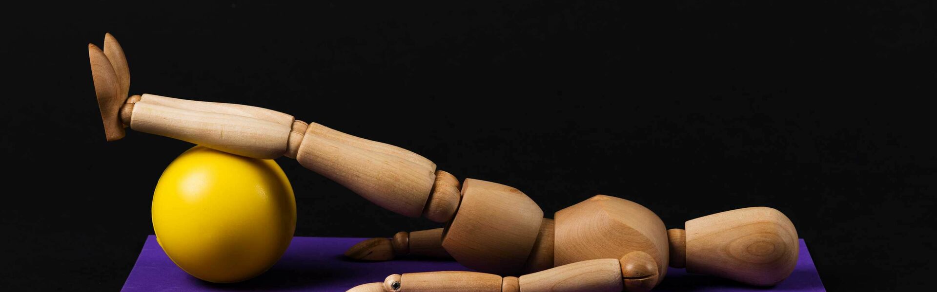 Symbolbild einer Holzfigur, die liegend eine Übung mit einem großen gelben Gymnastikball macht